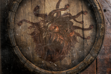 Un viejo barril sobre un suelo de madera desde una vista cenital. En el centro del barril, dibujado en la madera, se puede ver una bestia redondeada con unos tentáculos con ojos que le brotan de la cabeza, la bestia posee un solo ojo y una boca enorme con dientes afilados.